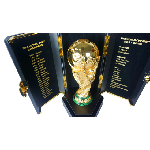 A gold FIFA World Cup trophy replica in a black case. The side panels list past champions and the host cities for the 2026 World Cup.