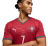 A man wears a red Puma Portugal soccer jersey with the number 7, featuring wavy patterns and green trim, posing with arms crossed against a black background.
