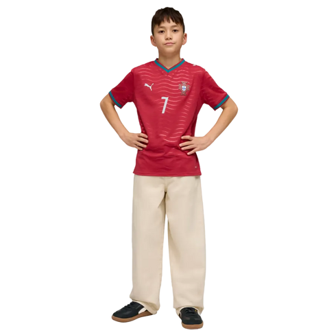 A boy posing in a red soccer jersey with the number 7, wide-leg cream trousers, and black sneakers against a black background.