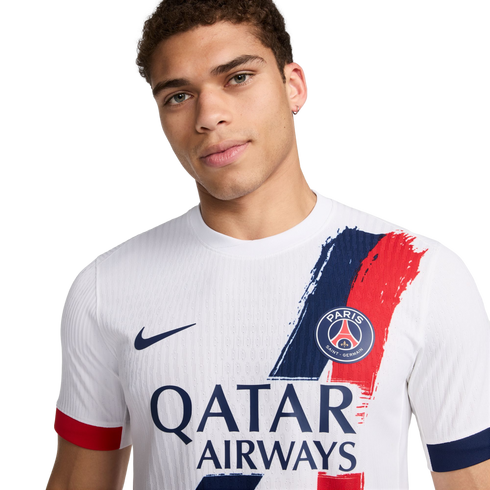 A man posing in a white Nike Paris Saint-Germain (PSG) away jersey featuring a blue and red brushstroke design and the Qatar Airways sponsor logo.