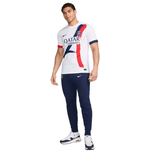 A male model wearing a white Paris Saint-Germain jersey with blue and red graphic details, navy Nike joggers, and white sneakers against a white background.