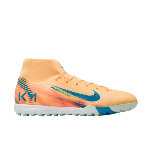 Side profile of a pale orange Nike Mercurial turf soccer shoe with teal accents and Kylian Mbappé's "KM" logo on the heel, set against a black background.