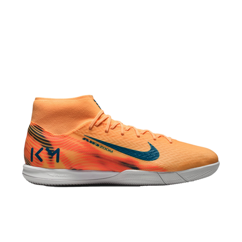 Side profile of an orange Nike soccer shoe with blue branding and a white sole. Features a sock-like ankle collar and "KM" logo on the heel.