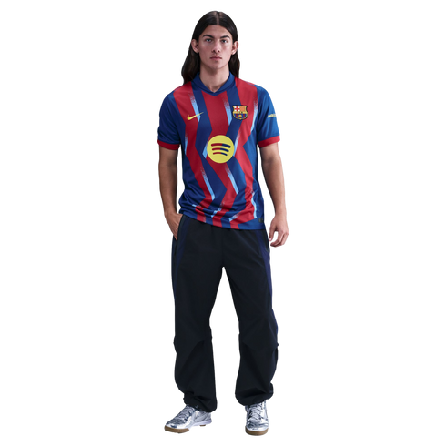 A young man with long hair wearing a red and blue zig-zag FC Barcelona jersey, dark track pants, and silver sneakers against a black background.