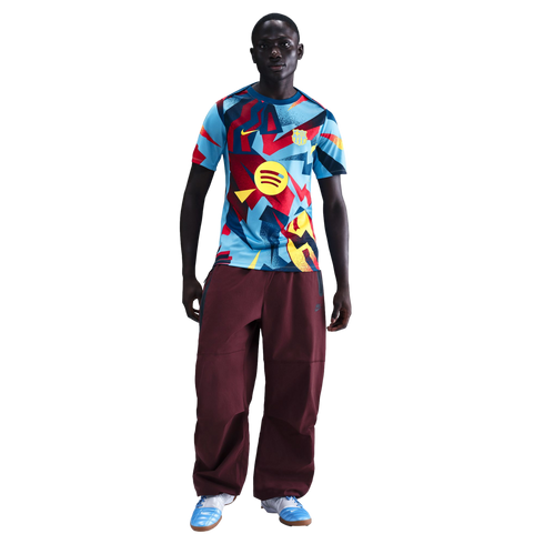Man wearing a patterned FC Barcelona jersey, maroon baggy pants, and light blue sneakers against a black background.