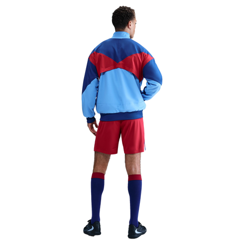 Back view of a man wearing a blue, light blue, and red color-blocked jacket, red shorts, and navy socks with red trim against a black background.