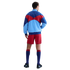 Back view of a man wearing a blue, light blue, and red color-blocked jacket, red shorts, and navy socks with red trim against a black background.