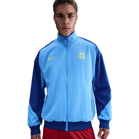 A man models a light and dark blue Nike FC Barcelona track jacket with a yellow club crest, posed against a solid black background.