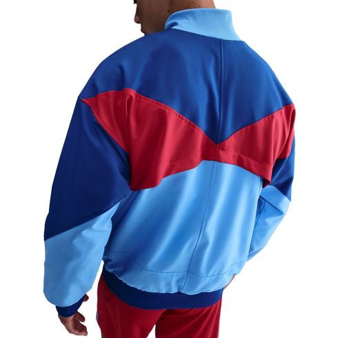 Rear view of a royal blue, light blue, and red color-blocked track jacket with geometric panels, worn with matching red shorts against a black background.