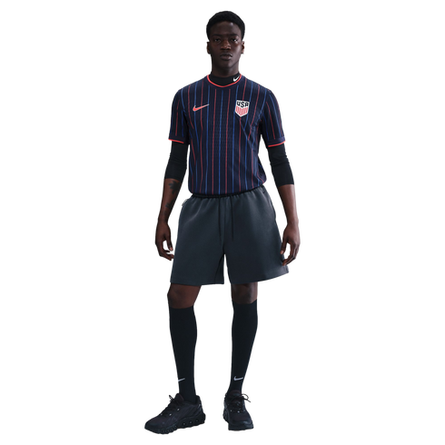 A man models a navy blue USA soccer jersey with red and blue pinstripes, charcoal gray shorts, black knee-high socks, and black sneakers.