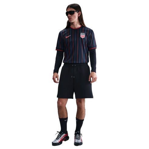 A person with long hair in sunglasses wearing a black pinstriped USA soccer jersey, black shorts, and Nike sneakers against a white background.