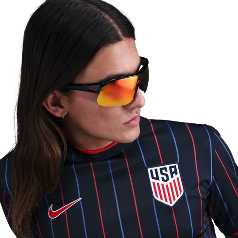 A person with long hair wearing a navy blue USA soccer jersey with thin red and blue stripes and orange reflective sunglasses.