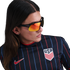 A person with long hair wearing a navy blue USA soccer jersey with thin red and blue stripes and orange reflective sunglasses.