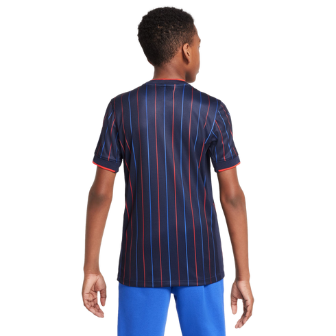 Rear view of a boy wearing a dark blue jersey with vertical red and blue stripes and bright blue pants against a white background.
