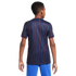 Rear view of a boy wearing a dark blue jersey with vertical red and blue stripes and bright blue pants against a white background.