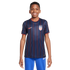 A smiling boy wearing a navy blue USA soccer jersey with red and blue pinstripes and blue sweatpants against a white background.