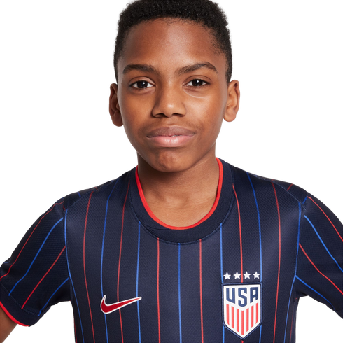 A young Black boy wearing a navy blue USA soccer jersey with red and blue stripes, a Nike swoosh, and the USA crest, posed against a white background.