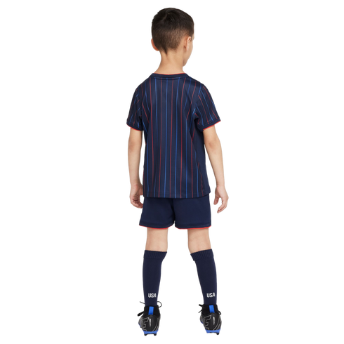 Rear view of a boy in a navy blue USA soccer kit with red and blue pinstripes, matching shorts with red trim, navy socks labeled "USA," and black soccer cleats.