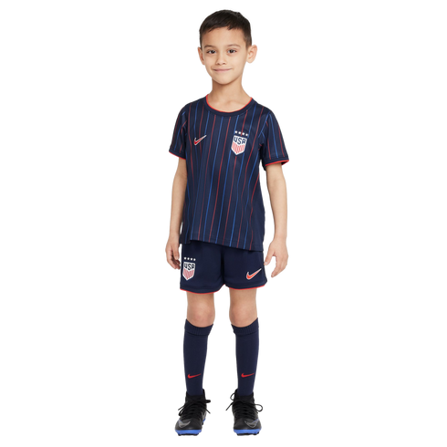 A young boy stands against a white background wearing a dark blue USA soccer kit with red and blue vertical stripes, matching shorts and socks, and black soccer cleats.
