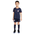 A young boy stands against a white background wearing a dark blue USA soccer kit with red and blue vertical stripes, matching shorts and socks, and black soccer cleats.