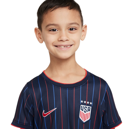 A smiling boy with a missing tooth wears a navy blue Nike USA soccer jersey with red and blue vertical stripes. White background.