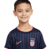A smiling boy with a missing tooth wears a navy blue Nike USA soccer jersey with red and blue vertical stripes. White background.