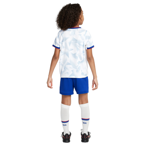 Back view of a child wearing a white USA soccer kit with geometric patterns, blue shorts, white socks with "USA" text, and black cleats.