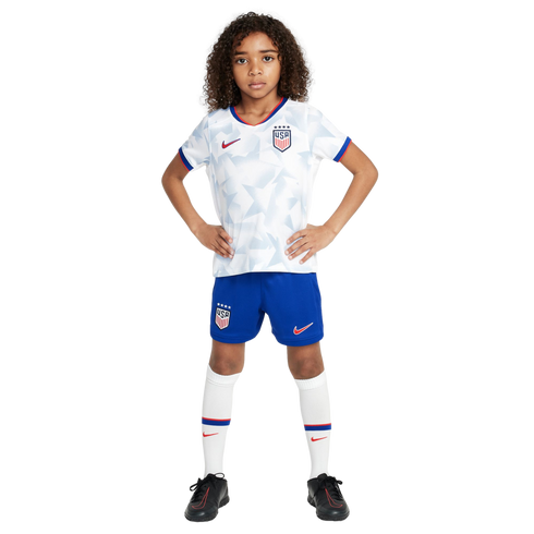 A young child in a white USA soccer uniform, blue shorts, and black cleats stands with hands on hips against a white background.