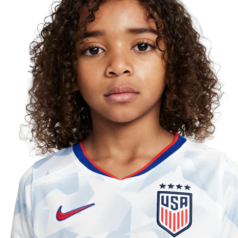A young child with curly brown hair wears a white USA soccer jersey, looking seriously at the camera against a plain white background.