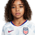 A young child with curly brown hair wears a white USA soccer jersey, looking seriously at the camera against a plain white background.