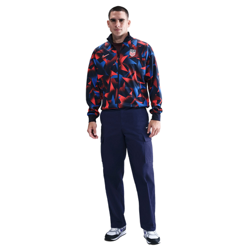 A man wears a red, blue, and black patterned Nike USA tracksuit jacket, navy cargo pants, and white sneakers against a white background.