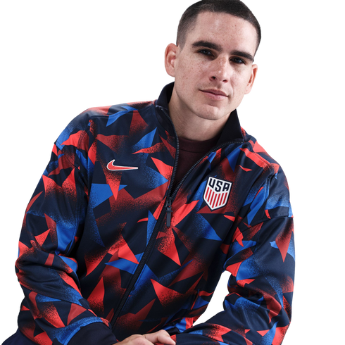 A man with freckles wears a red and blue geometric Nike USA national soccer team jacket against a white background.