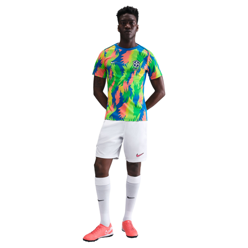 A Black man models a colorful Nike Brazil soccer kit with an abstract pattern jersey, white shorts and socks, and pink cleats against a white background.