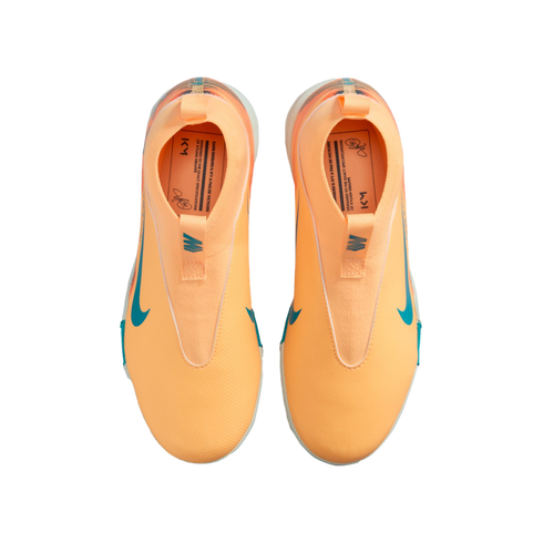 Top-down view of bright orange laceless Nike soccer shoes with teal accents and "KM" branding on the insoles, set against a black background.