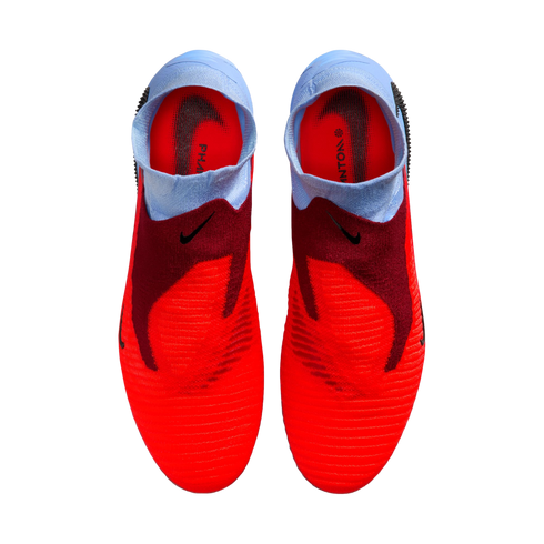 A top-down view of a pair of red and light blue laceless Nike Phantom football boots against a solid black background.