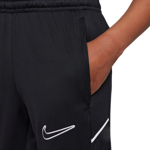 Close-up of black Nike athletic pants with a hand in the side pocket. Features a white embroidered swoosh logo and a diagonal white stripe.