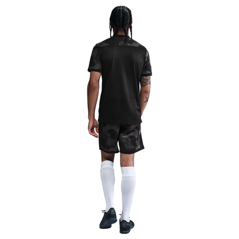 Back view of a man with braided hair wearing a black soccer jersey, patterned shorts, tall white socks, and black cleats.