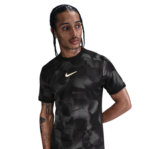 A man with braided hair wears a black patterned Nike t-shirt and looks to the side against a white background.