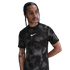 A man with braided hair wears a black patterned Nike t-shirt and looks to the side against a white background.