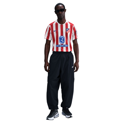 A man stands wearing a red and white striped Atlético de Madrid jersey, black Nike track pants, and black sunglasses against a white background.