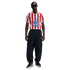 A man stands wearing a red and white striped Atlético de Madrid jersey, black Nike track pants, and black sunglasses against a white background.