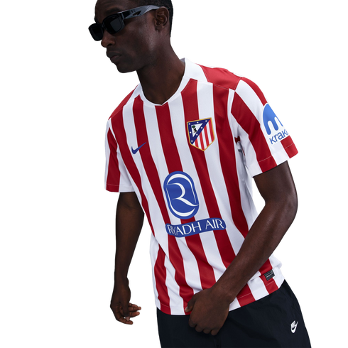 A person in sunglasses wearing a red and white striped Atletico Madrid soccer jersey with Riyadh Air and Kraken sponsors, standing against a white background.