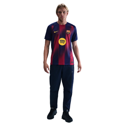 A blonde man wearing an FC Barcelona football jersey, navy track pants, and black trainers, standing against a solid black background.