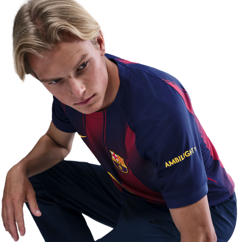 A young man with blonde hair squats and looks intently over his shoulder while wearing an FC Barcelona jersey against a solid black background.