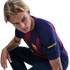 A young man with blonde hair squats and looks intently over his shoulder while wearing an FC Barcelona jersey against a solid black background.