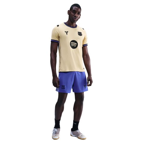 A man modeling a gold FC Barcelona x Kobe Bryant jersey with purple shorts and black socks against a black background.