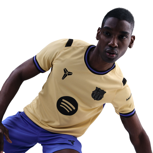 A man in a tan FC Barcelona football jersey featuring Kobe Bryant’s "Sheath" logo and a Spotify logo, wearing blue shorts against a black background.