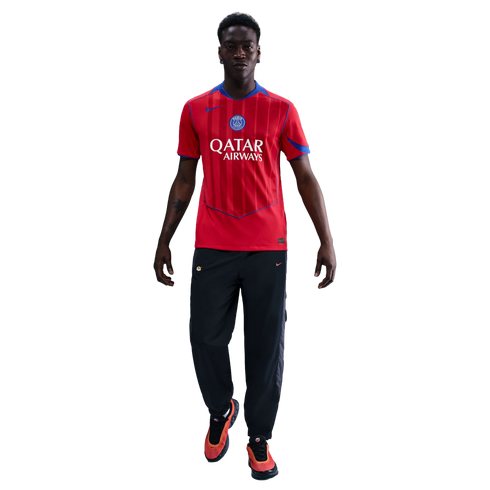 A man modeling a red Paris Saint-Germain soccer jersey, black track pants, and orange sneakers against a black background.