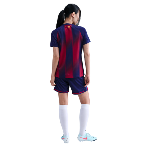 Rear view of a woman in a red and blue striped soccer kit, white socks, and light blue cleats against a black background.