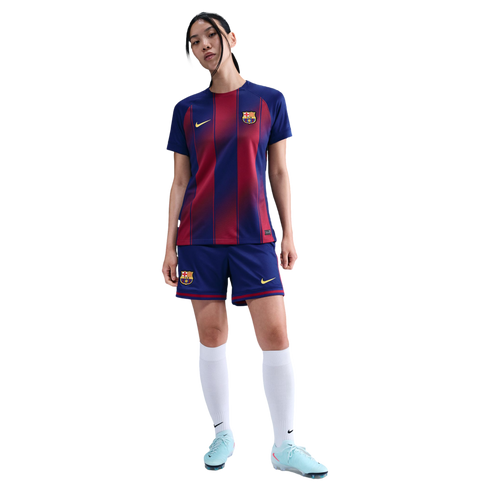 A woman wears an FC Barcelona soccer kit with a blue and red striped jersey, blue shorts, white socks, and light blue cleats, standing against a black background.